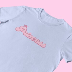 May include: A light blue t-shirt with a pink graphic of the word "Princess" with a crown above it.