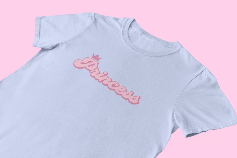May include: A light blue t-shirt with a pink graphic of the word "Princess" with a crown above it.