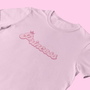 May include: A light pink t-shirt with the word "Princess" in a pink retro font with a crown above the word.