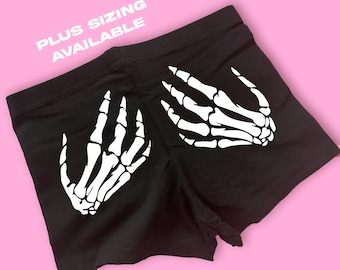 May include: Black shorts with white skeleton hand graphics on the sides. The text "PLUS SIZING AVAILABLE" is printed in white on the top left. The shorts are displayed on a pink background.