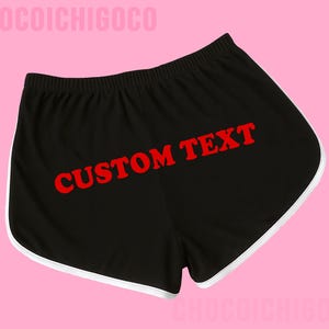 May include: Black athletic-style shorts featuring a white trim along the curved leg openings and an elastic waistband. The centre of the shorts displays the words CUSTOM TEXT printed in a bold, bright red font against the black fabric background.