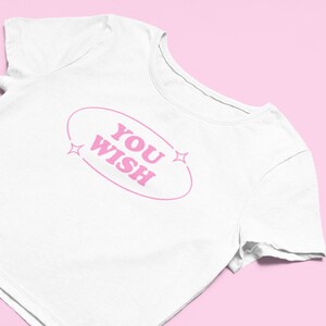 May include: White crop top with pink text that reads "YOU WISH" inside an oval with two pink stars on either side.