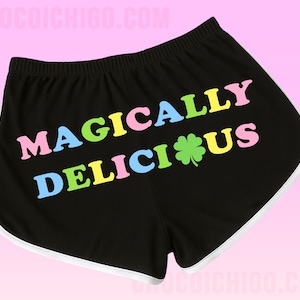 Op de afbeelding: Zwarte shorts met witte rand en de tekst "MAGICAL DELICIOUS" in kleurrijke letters, met een groene klaver. De shorts hebben een elastische tailleband.