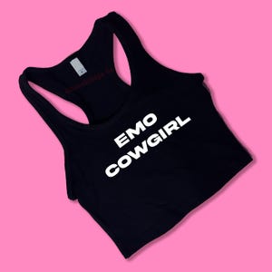 May include: Black racerback tank top with the words "EMO COWGIRL" in white block letters. The tank top has a scoop neck and a racerback design. The brand name "Chocolate Co" is printed in red on the back.