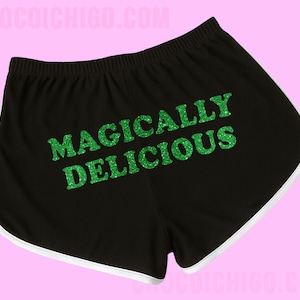 Könnte beinhalten: Schwarze Sporthosen mit weißem Besatz und dem Text "MAGICALY DELICIOUS" in grünem Glitzer. Die Shorts haben einen elastischen Bund und eine bequeme Passform.