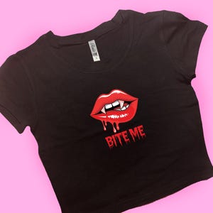 May include: Black crop top featuring a red graphic of vampire lips with fangs and dripping blood, and the text "BITE ME". The top has a scoop neck and short sleeves.