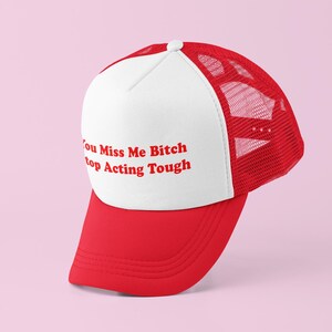 Peut inclure: Casquette de camionneur rouge et blanche avec le texte "You Miss Me Bitch Stop Acting Tough" en rouge. La casquette a une visière rouge et un panneau avant blanc avec le texte. L'arrière de la casquette est en mesh rouge.