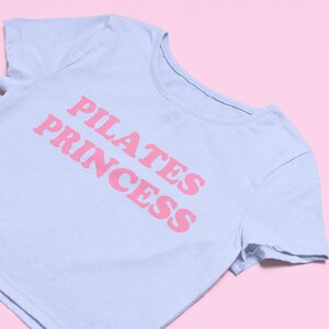 Puede incluir: Una camiseta corta de color azul claro con el texto "PILATES PRINCESS" impreso en rosa.