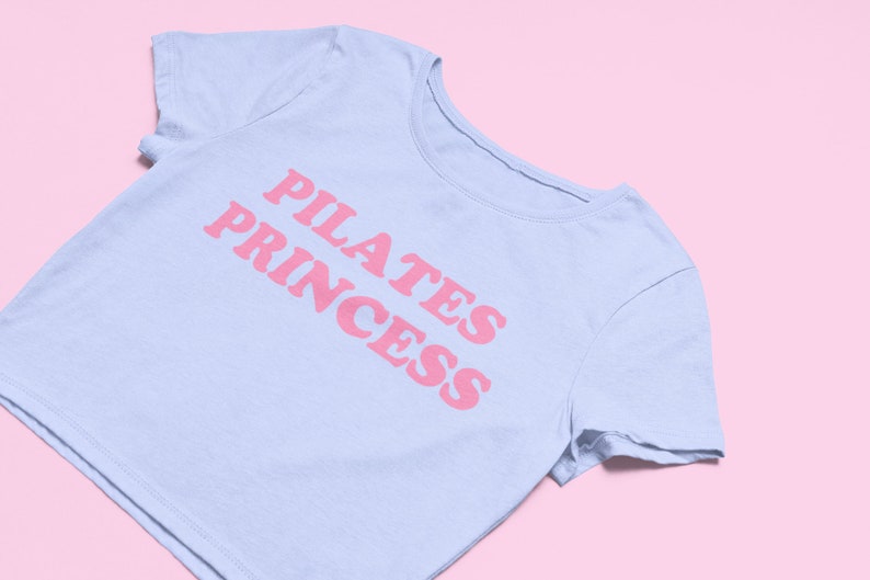 Puede incluir: Una camiseta corta de color azul claro con el texto "PILATES PRINCESS" impreso en rosa.