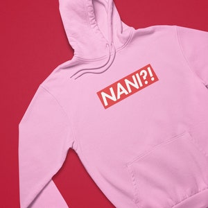 K&ouml;nnte beinhalten: Hellrosa Kapuzenpullover mit Kordelzug und Vordertasche. Der Schriftzug "NANI?!" ist in wei&szlig;en Buchstaben in einem roten Rechteck auf der Vorderseite aufgedruckt.