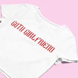 May include: White t-shirt with the red text "GOTH GIRLFRIEND" in a gothic font. The shirt is displayed on a pink background. The t-shirt is a casual wear item.