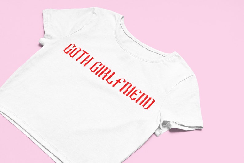 May include: White t-shirt with the red text "GOTH GIRLFRIEND" in a gothic font. The shirt is displayed on a pink background. The t-shirt is a casual wear item.