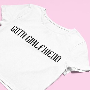 May include: White t-shirt with the text "GOTH GIRLFRIEND" in a black gothic font. The shirt is angled, showing the neckline and sleeve, set against a pink background.