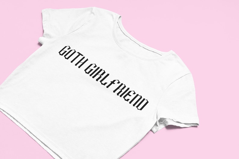 May include: White t-shirt with the text "GOTH GIRLFRIEND" in a black gothic font. The shirt is angled, showing the neckline and sleeve, set against a pink background.