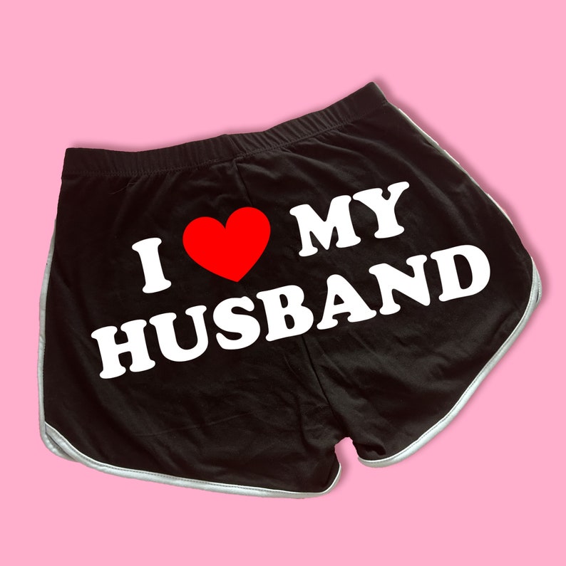 May include: Black athletic shorts with a white trim and the text "I love my husband" in white letters with a red heart.