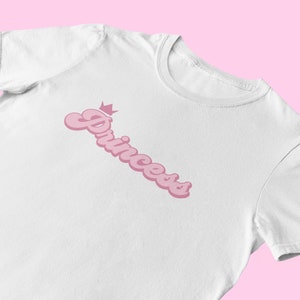 May include: White t-shirt with a pink "Princess" graphic with a crown above the word.