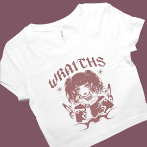 May include: White t-shirt with a crew neck and short sleeves. The shirt features a brown graphic of a manga-style girl with the word "WRAITHS" above her. The graphic includes stars and decorative elements.