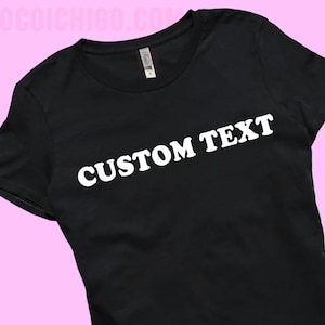 May include: Black crew neck t-shirt with the words "CUSTOM TEXT" printed in white across the front. The shirt is displayed on a pink background. The text is in a bold, sans-serif font.