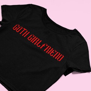 May include: Black t-shirt with the red text "GOTH GIRL FRIEND" in a gothic font. The shirt is displayed on a pink background. The t-shirt is a crew neck style.