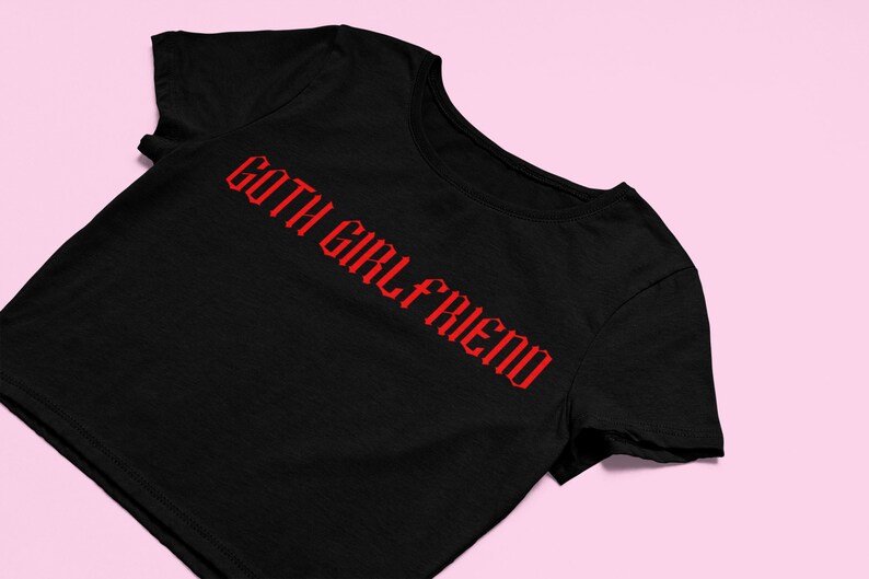May include: Black t-shirt with the red text "GOTH GIRL FRIEND" in a gothic font. The shirt is displayed on a pink background. The t-shirt is a crew neck style.