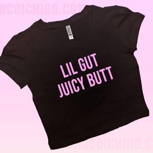May include: Black crop top with the text "LIL GUT JUICY BUTT" in pink. The shirt has short sleeves and a crew neck. The background is a gradient of pink.