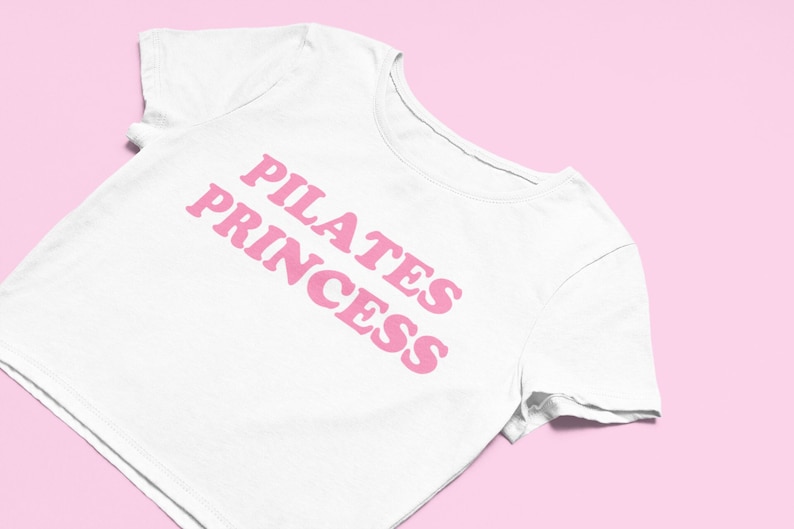 Puede incluir: Camiseta blanca recortada con texto rosa que dice "Pilates Princess".