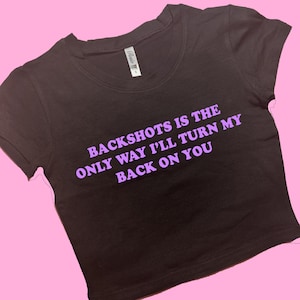 May include: A black crop top with the text "BACKSHOTS IS THE ONLY WAY I'LL TURN MY BACK ON YOU" in purple. The shirt has short sleeves and a round neckline. The background is pink.