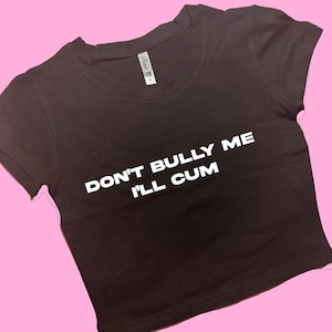 May include: A dark brown crop top with the text "DON'T BULLY ME I'LL CUM" in white block letters. The shirt has short sleeves and a crew neck. The background is a solid pink color.