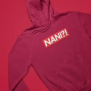 K&ouml;nnte beinhalten: Ein weinroter Kapuzenpullover mit einer Vordertasche und einer Kapuze. Der Kapuzenpullover zeigt das Wort "NANI?!" in wei&szlig;em Text in einem roten Rechteck.