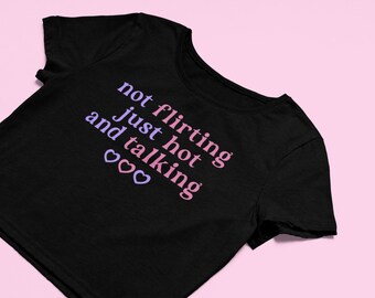 May include: Black t-shirt with the text "not flirting just hot and talking" in pink and purple. The shirt has two heart symbols below the text. The background is a solid light pink.