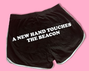 May include: Black athletic shorts with white trim and the text "A NEW HAND TOUCHES THE BEACON" in white block letters. The shorts have an elastic waistband and a retro style.