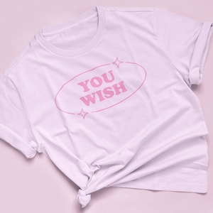 May include: White t-shirt with rolled sleeves and a knotted hem. The shirt features the text "YOU WISH" in pink, inside a pink oval with star accents. The shirt is displayed on a pink surface.