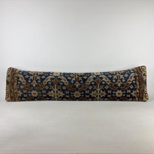 May include: A long, rectangular bolster pillow featuring a dark blue, beige, and brown intricate floral pattern. The design displays a repeating symmetrical motif across the entire surface, with decorative borders on both ends of the pillow.