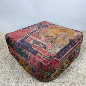 Pode incluir: Um pufe quadrado estampado com um design de tapete vintage. O pufe apresenta uma mistura de cores, incluindo vermelho, laranja, azul e verde. O design parece ser um padrão geométrico tradicional. O pufe é colocado sobre um tapete branco e felpudo.