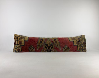 May include: A long, rectangular bolster pillow featuring a woven textile cover with a deep red central field, tan accents, and dark blue floral motifs. The fabric displays a dense, intricate pattern across the entire surface of the cushion.