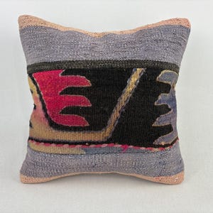 May include: Square decorative pillow with a woven design. The pillow features a central black panel with red, yellow, and blue geometric shapes. The edges are a light purple with a pink border, creating a textured look.