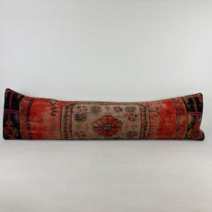 May include: A long, rectangular pillow with a distressed, vintage look. The pillow features a red and beige floral pattern with black accents on the ends. The pillow is approximately 36 inches long.