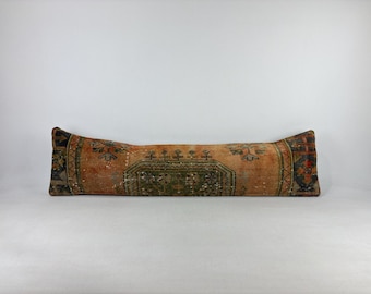 May include: A long, rectangular pillow with an aged, distressed appearance. The pillow features a central design in shades of green and brown, with orange and black accents. The pillow's design suggests a vintage or antique aesthetic, suitable for home decor.