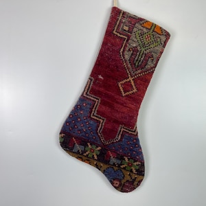 May include: A Christmas stocking with a red background and geometric patterns in blue, green, and gold. The stocking is made of a woven material and has a loop for hanging.