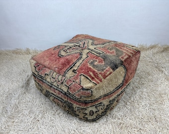May include: A square floor pouf crafted from woven textile featuring a faded red, beige, and black geometric pattern. The low-profile seat has a textured, knotted surface with visible stitching along the edges, creating a structured, boxy shape.