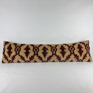 May include: A long, rectangular lumbar pillow featuring a repeating damask-style pattern in deep burgundy red set against a textured, light beige background. The fabric has a woven appearance with a consistent, symmetrical design across the surface.