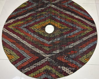 Pode incluir: Uma saia de árvore circular com um padrão geométrico tecido em tons de castanho escuro, vermelho profundo, verde azeitona, laranja e cinzento. O tecido tem uma aparência texturizada com um recorte central circular e uma fenda reta.