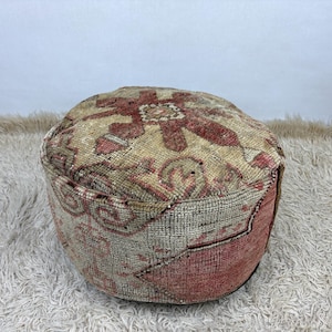 Puede incluir: Un puf de suelo redondo hecho de tejido con un patrón descolorido y desgastado en tonos rojo apagado, beige y crema. La superficie superior muestra un motivo floral geométrico, mientras que los lados presentan un aspecto texturizado.