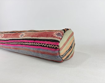May include: A cylindrical bolster pillow featuring a woven textile cover with horizontal stripes in shades of red, pink, gray, and black. The fabric displays small geometric diamond patterns and the circular end panel has multi-colored stripes.