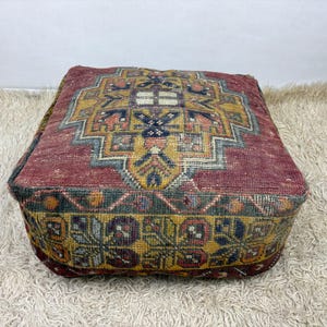 May include: A square floor pouf featuring a woven textile surface with a deep red base color. The top displays a large, intricate geometric medallion in shades of mustard yellow, navy blue, and cream. The sides feature a repeating floral pattern.