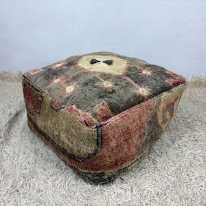 May include: A square floor pouf featuring a thick, textured wool pile with a multi-colored geometric pattern in shades of dark brown, muted red, and cream. The surface displays a central diamond motif surrounded by smaller star-like shapes.