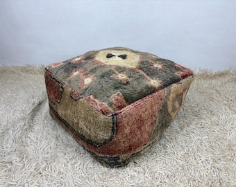 Peut inclure: Un pouf de sol carré doté d'un poil en laine épais et texturé avec un motif géométrique multicolore dans des tons brun foncé, rouge sourd et crème. La surface présente un motif central en losange entouré de petites formes étoilées.