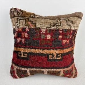 May include: A square, textured pillow with a traditional rug design. The pillow features a color palette of red, brown, beige, and touches of pink. The design includes geometric patterns and floral motifs, creating a cozy, decorative accent.