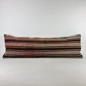 May include: A long, rectangular lumbar pillow featuring a woven textile cover with horizontal stripes in shades of black, orange, grey, and muted red. The fabric displays a textured, multi-colored pattern across the entire surface of the cushion.