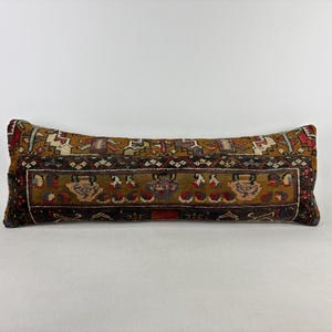 May include: A long rectangular lumbar pillow featuring a woven textile cover with a complex geometric and floral pattern in shades of brown, dark red, blue, and cream. The design includes a central horizontal band with repeating motifs.